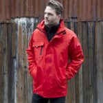 Mann trägt eine leuchtend rote 3-in-1 Transitjacke vor einer hölzernen Wand.