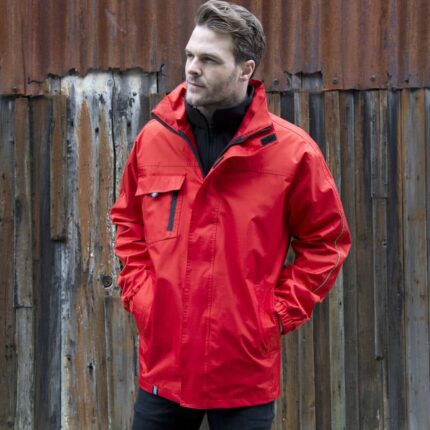 Mann trägt eine leuchtend rote 3-in-1 Transitjacke vor einer hölzernen Wand.