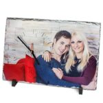 Personalisierte Wanduhr aus Schiefer mit Fotodruck eines Paares, montiert auf schwarzem Standfuß mit sichtbaren Uhrzeigern.