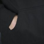 Close-up of a person’s hand inside the front pocket of a black Meenzer Fastnachtshoodie, showing the ribbed sleeve cuff and fabric texture